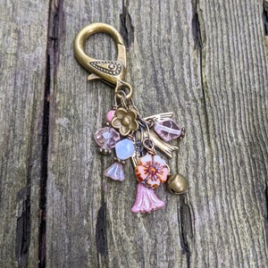 Pu&ograve; includere: Un ciondolo decorativo in stile vintage con chiusura in ottone e vari elementi. Include perline rosa e trasparenti, piccoli fiori e un campanello. La palette di colori presenta tonalit&agrave; ottone, rosa e azzurro.
