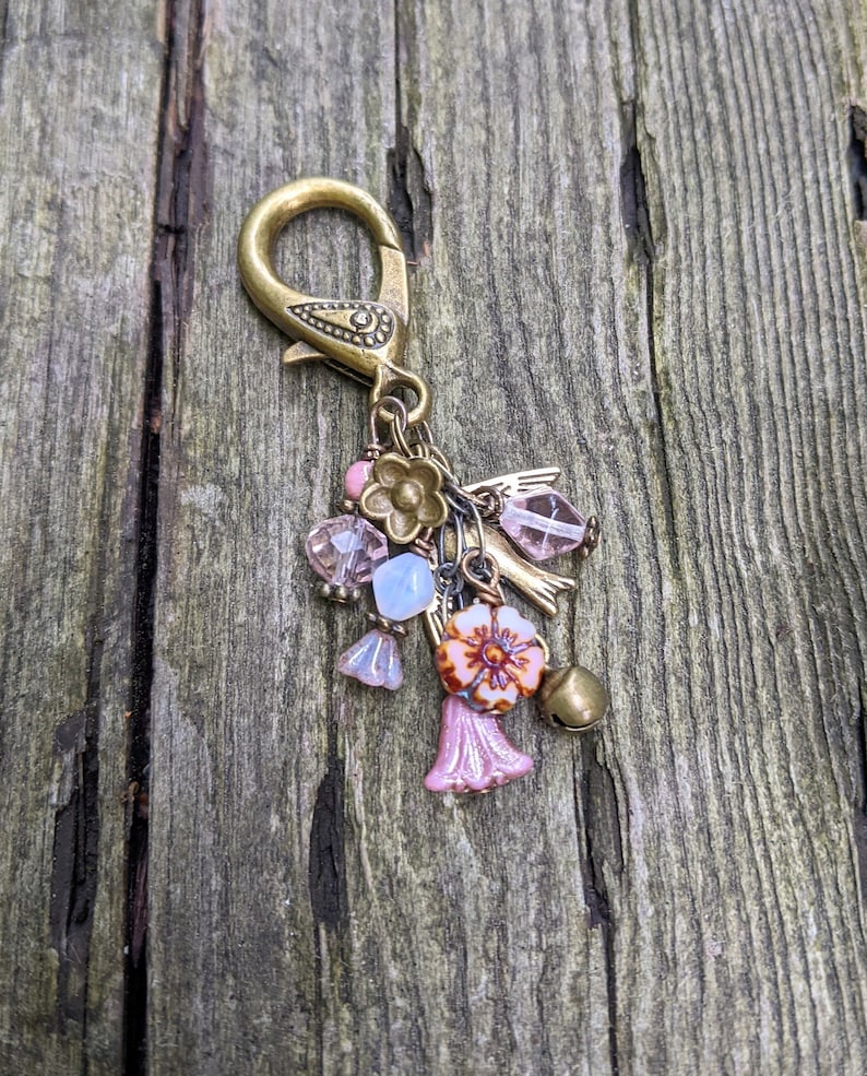 Pu&ograve; includere: Un ciondolo decorativo in stile vintage con chiusura in ottone e vari elementi. Include perline rosa e trasparenti, piccoli fiori e un campanello. La palette di colori presenta tonalit&agrave; ottone, rosa e azzurro.