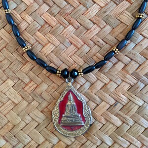 May include: A black beaded necklace with a gold pendant featuring a seated Buddha figure against a red background. The pendant is teardrop-shaped with ornate detailing. The necklace is displayed on a woven surface.