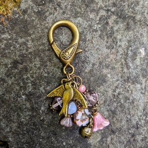 Pu&ograve; includere: Un ciondolo color bronzo con un ciondolo a forma di uccello e un moschettone. Il ciondolo presenta un gruppo di perline rosa, viola e bianche, insieme a una piccola campana e perline a forma di fiore. Lo sfondo &egrave; una pietra grigia strutturata.