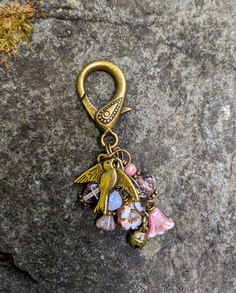 Pu&ograve; includere: Un ciondolo color bronzo con un ciondolo a forma di uccello e un moschettone. Il ciondolo presenta un gruppo di perline rosa, viola e bianche, insieme a una piccola campana e perline a forma di fiore. Lo sfondo &egrave; una pietra grigia strutturata.