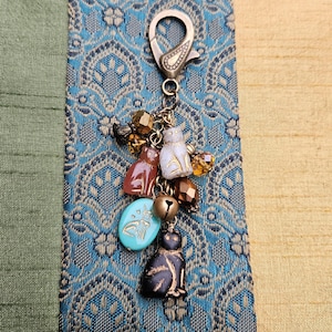 May include: A silver-tone charm with a lobster clasp, featuring cat-shaped charms in various colors, including brown, white, and black. The charm also includes a turquoise-colored oval bead and a small gold bell, with amber-colored beads.