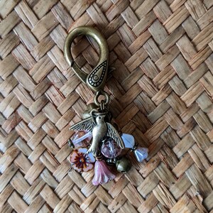 Pu&ograve; includere: Ciondolo portachiavi color ottone antico con un uccello dettagliato con le ali spiegate, circondato da perline rosa e trasparenti e piccoli ciondoli a forma di fiore rosa. Il ciondolo &egrave; attaccato a un grande fermaglio.