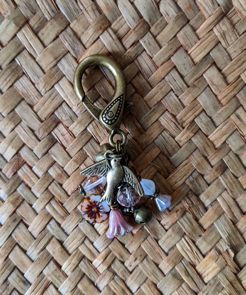 Pu&ograve; includere: Ciondolo portachiavi color ottone antico con un uccello dettagliato con le ali spiegate, circondato da perline rosa e trasparenti e piccoli ciondoli a forma di fiore rosa. Il ciondolo &egrave; attaccato a un grande fermaglio.