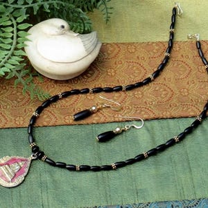 May include: A black beaded necklace and matching earrings set with gold accents. The necklace features a pendant with pink and gold detailing. The jewelry is displayed on a green and brown patterned surface.