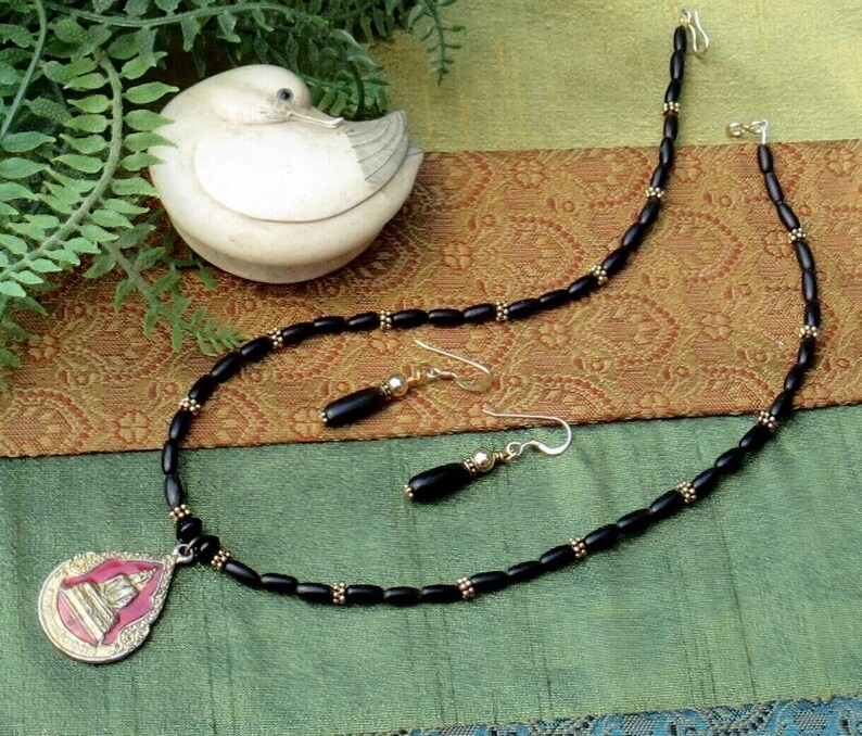 May include: A black beaded necklace and matching earrings set with gold accents. The necklace features a pendant with pink and gold detailing. The jewelry is displayed on a green and brown patterned surface.