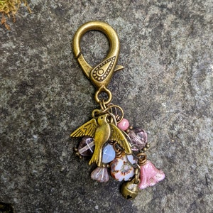 Pu&ograve; includere: Un ciondolo decorativo color bronzo con un ciondolo a forma di uccello, perline rosa e trasparenti e una piccola campana. Il ciondolo &egrave; attaccato a una chiusura color bronzo. Il ciondolo &egrave; posto su uno sfondo di pietra grigia.