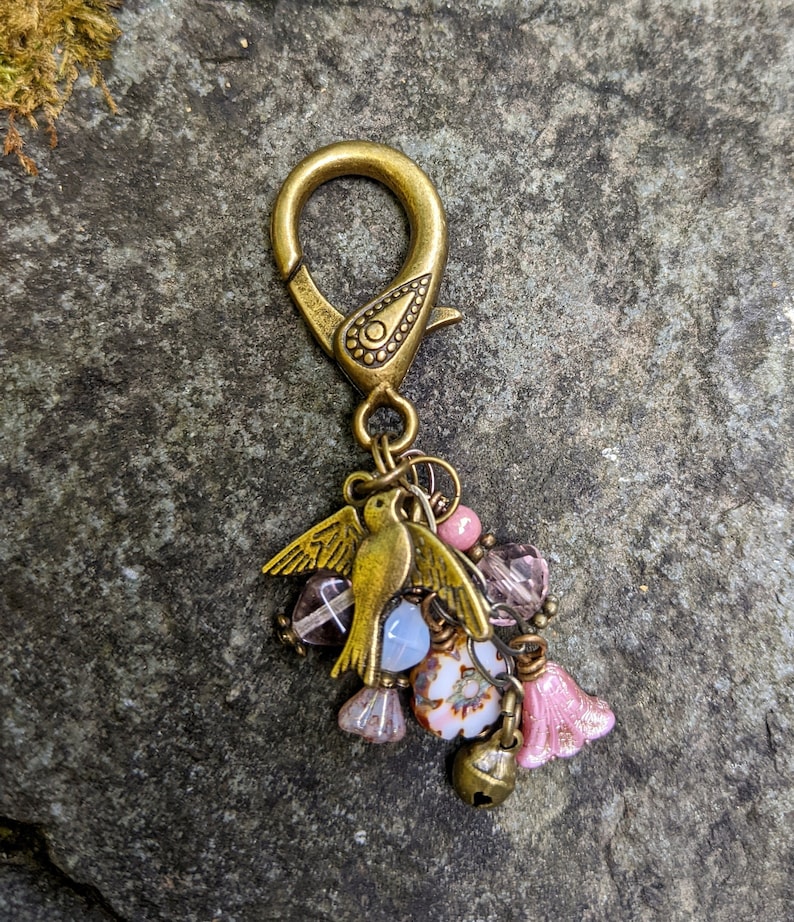 Pu&ograve; includere: Un ciondolo decorativo color bronzo con un ciondolo a forma di uccello, perline rosa e trasparenti e una piccola campana. Il ciondolo &egrave; attaccato a una chiusura color bronzo. Il ciondolo &egrave; posto su uno sfondo di pietra grigia.