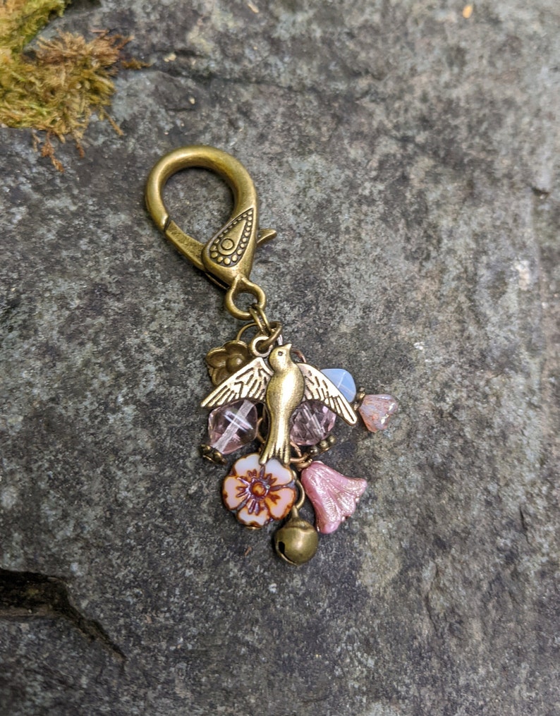Pu&ograve; includere: Un ciondolo color bronzo con un uccello, fiori e perline. Il ciondolo ha un moschettone e presenta un piccolo uccello con le ali spiegate, fiori rosa e bianchi e varie perline. Una piccola campana &egrave; appesa sotto i fiori.