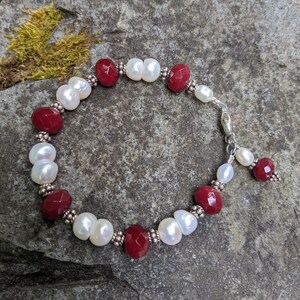 May include: A beaded bracelet featuring alternating red gemstone beads and white pearl beads. The bracelet is strung on a silver chain and has a silver clasp. The bracelet is displayed on a gray stone surface.