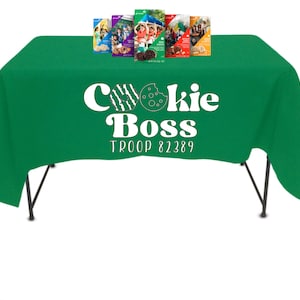 May include: A green tablecloth with the words "Cookie Boss Troop 82389" in white. Several boxes of cookies are displayed on the table. The table has black legs and is set against a white background.