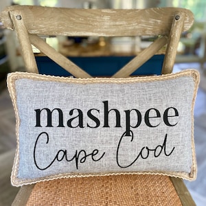 以下が含まれることがあります： 「mashpee Cape Cod」の文字が黒い筆記体で書かれた長方形のグレーのクッション。クッションは薄いベージュのトリムで、編み込まれた座面のある木製の椅子の上に置かれています。