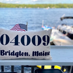 May include: A white pillow with the text "04009 Bridgton, Maine" in black lettering. The pillow is sitting on a wooden surface with a blurry background of a lake and boats.