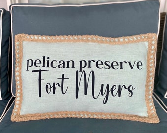 May include: A light blue throw pillow with a burlap trim and the text "pelican preserve Fort Myers" in black.