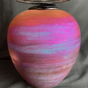May include: A decorative urn with a gradient of colors, transitioning from red to purple and blue. The urn has a dark metal lid with a small green accent and a curved handle. The overall shape is rounded and bulbous.