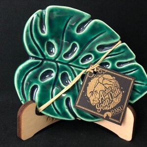 May include: A green ceramic soap dish shaped like a monstera leaf. The dish is tied with a natural fiber string and includes a tag that reads "Aloha Soap Dish". The dish is displayed on a wooden stand.