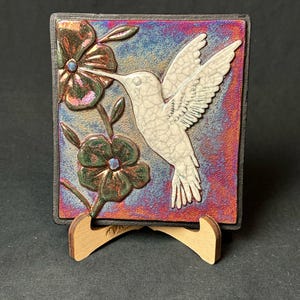 May include: A square ceramic tile titled Hummingbird 1, featuring a white crackle-glazed hummingbird in flight toward two dark green flowers. The background displays a vibrant, iridescent gradient of blue, purple, and copper tones on a wooden stand.
