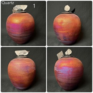 May include: A rounded ceramic vessel with a textured, iridescent finish in shades of deep purple, magenta, and orange. The lid is black and topped with a clear, raw quartz crystal point. The word Quartz and the number 1 appear in the top corner.