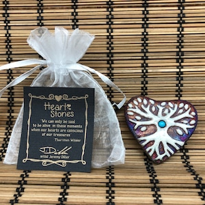 May include: A heart-shaped ceramic stone with a white tree of life design and a turquoise gemstone in the center. The stone is on a woven mat with a small white organza bag containing a card that reads "Heart Stones" and a quote by Thornton Wilder.