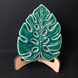 May include: A ceramic leaf-shaped dish in a vibrant green hue, featuring a glossy finish and intricate vein detailing. The dish is mounted on a small, wooden stand, creating a decorative display piece. The design is reminiscent of a Monstera plant.