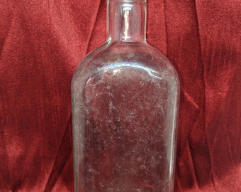 May include: A clear glass bottle with a rounded bottom and a straight neck. The bottle is empty and has a slightly textured surface.