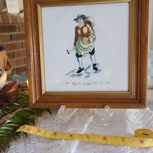 May include: A framed illustration of a man in a green and brown outfit carrying a large bag on his back. The illustration is on a white background with a black and white checkered floor. The text below the illustration reads "A Serving Foot or an Irish Foot to Mend.  Giles of London."