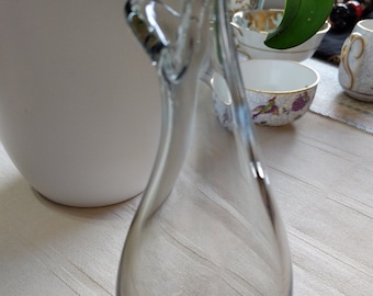 May include: A clear glass vase with a unique, curved design. The vase is shaped like a pear and has a wide opening at the top. It is sitting on a white tablecloth.
