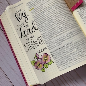May include: A close-up of an open Bible with a hand-lettered quote, "The joy of the Lord is my strength." The quote is written in black ink with a decorative flourish. A watercolor painting of a purple pansy flower is in the bottom right corner of the image.