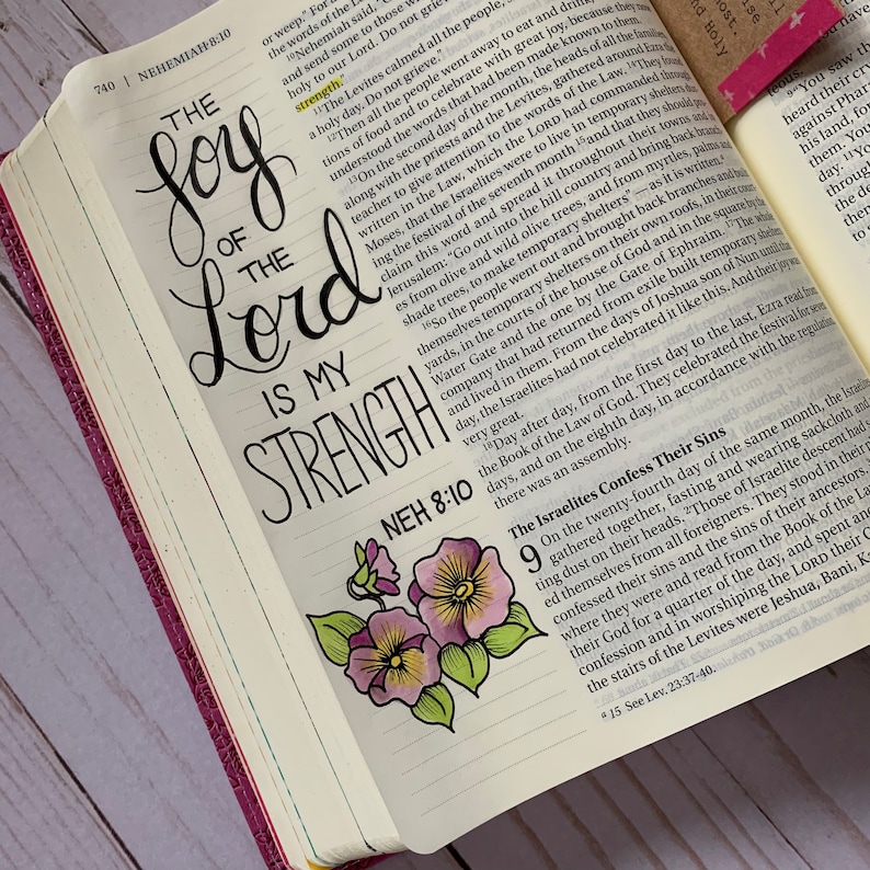 May include: A close-up of an open Bible with a hand-lettered quote, "The joy of the Lord is my strength." The quote is written in black ink with a decorative flourish. A watercolor painting of a purple pansy flower is in the bottom right corner of the image.