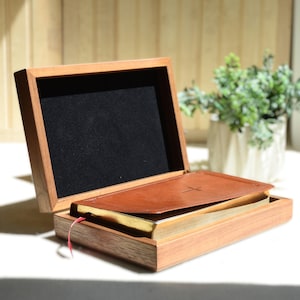 May include: A wooden box with a hinged lid, open to reveal a brown leather-bound book with a cross design. The box is made of light-colored wood and has a black velvet lining. A red ribbon bookmark is visible.