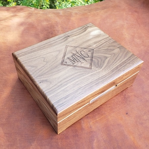 May include: A rectangular wooden box with a hinged lid. The box is made of light brown wood with a visible grain pattern. The lid features a diamond-shaped engraving with the letters "MNG".