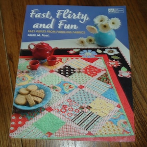 May include: A book titled "Fast, flirty, and fun: Easy quilts from fabulous fabrics" by Sarah M. Bisek. The cover features a colorful patchwork quilt with a red teapot and cookies on a wooden table.