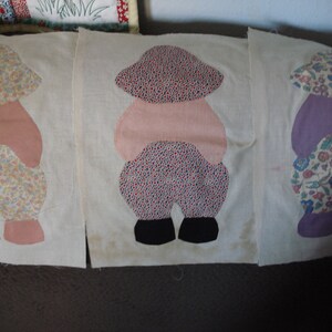May include: Three fabric squares with appliqued figures of people wearing hats and dresses. The figures are made of different patterned fabrics and are sewn onto a white background.