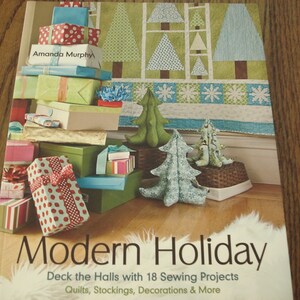 May include: A book titled "Modern Holiday: Deck the Halls with 18 Sewing Projects" with a colorful quilt design on the cover. The book features a stack of wrapped gifts with red and white polka dot paper, green and white striped paper, and a blue and white striped paper.