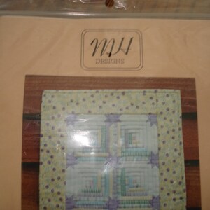 May include: A craft kit featuring a quilt design. The quilt pattern is visible through the clear packaging. The quilt design has a light green border with purple polka dots and stars. The center of the quilt has a log cabin design. The packaging has the text "MH DESIGNS".