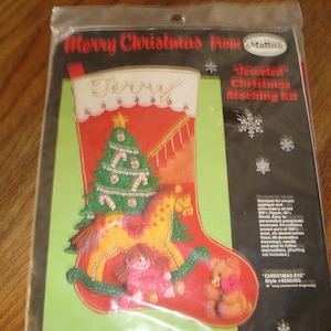 May include: A Christmas stocking kit with a red background, featuring a green Christmas tree, a rocking horse, and a doll. The text "Merry Christmas from Malina" and "Terry" are visible. The kit is 18 inches long.