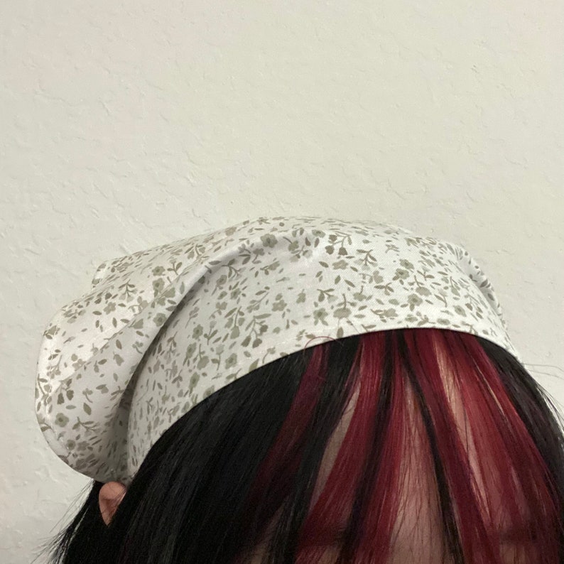 Pu&ograve; includere: Un bandana bianco con un piccolo motivo floreale verde. Il bandana &egrave; legato intorno alla testa di una persona con i capelli scuri e riflessi rossi.