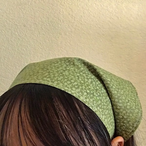 Pu&ograve; includere: Un bandana verde con un piccolo motivo floreale bianco. Il bandana &egrave; legato intorno alla testa di una persona.