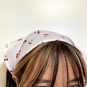 Pu&ograve; includere: Un bandana bianco con una stampa di ciliegie rosse. Il bandana &egrave; legato intorno alla testa di una persona con capelli castani e biondi.