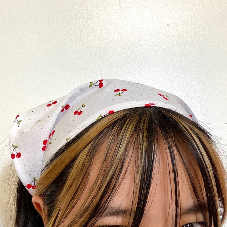 Pu&ograve; includere: Un bandana bianco con una stampa di ciliegie rosse. Il bandana &egrave; legato intorno alla testa di una persona con capelli castani e biondi.