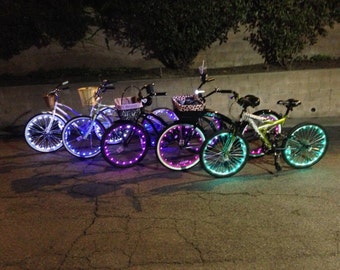 May include: Four bicycles with colorful LED lights on the wheels. The bikes are parked on a paved surface. The bikes have baskets and are decorated with various accessories.