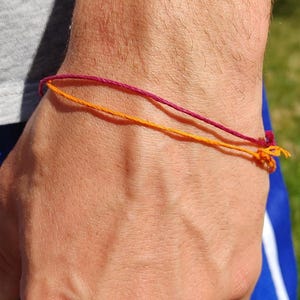May include: A close-up of a wrist adorned with two thin string bracelets. One bracelet is a deep magenta color, and the other is a vibrant orange. The bracelets are knotted at the ends.