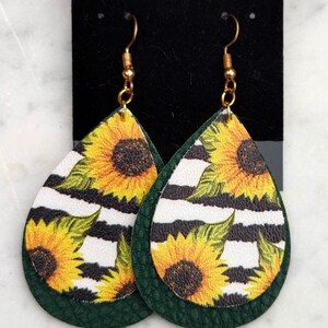 May include: Teardrop-shaped earrings featuring a sunflower design with yellow petals and a dark brown center, set against a black and white striped background. The earrings have a dark green backing and gold-colored hooks.