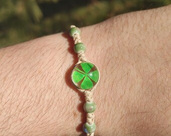May include: A handmade bracelet featuring a central charm with a four-leaf clover design. The charm is surrounded by a clear border and is connected to the bracelet with a beige woven cord. Green beads are interspersed along the cord.