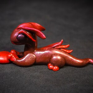 Op de afbeelding: Een klein, rood en bruin drakenbeeldje met een glanzende afwerking. De draak ligt op zijn buik met zijn kop naar de zijkant gedraaid. Hij heeft een rood hart in zijn voorpoten.