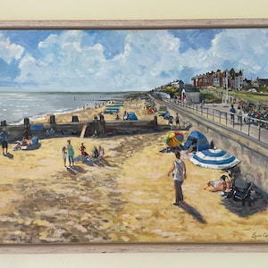 May include: Oil painting of a beach scene with people, umbrellas, and buildings. The artwork depicts a sandy beach with a promenade, blue sky, and fluffy white clouds. The painting is framed in a light-coloured wooden frame.