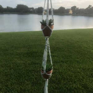 May include: A white macrame plant hanger with two small terracotta pots and green plants hanging from it. The hanger is hanging in front of a green grassy lawn and a lake in the background.