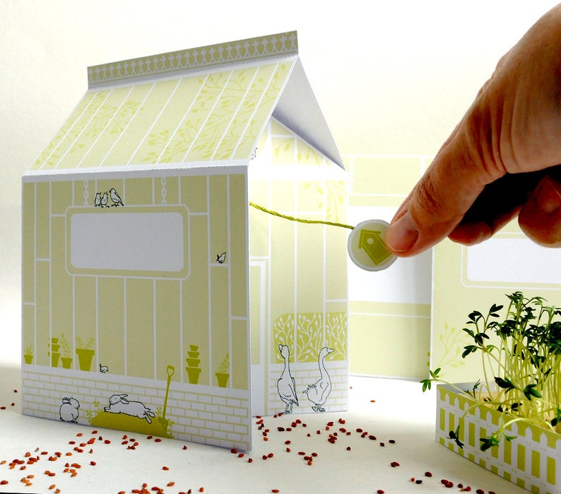 May include: A papercraft model of a greenhouse with a green roof, white walls, and a yellow interior. The greenhouse has a small door, a window, and a birdhouse. There are two birds on the roof of the greenhouse. The greenhouse is sitting on a white surface with red sprinkles.