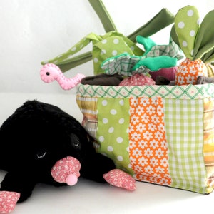 May include: A patchwork fabric basket filled with plush vegetables and a pink worm toy. A black plush mole with pink floral accents is in the foreground. The basket has green, orange, and yellow patterned fabric.