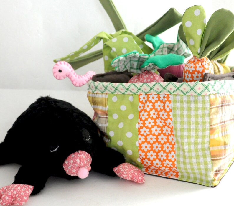 May include: A patchwork fabric basket filled with plush vegetables and a pink worm toy. A black plush mole with pink floral accents is in the foreground. The basket has green, orange, and yellow patterned fabric.